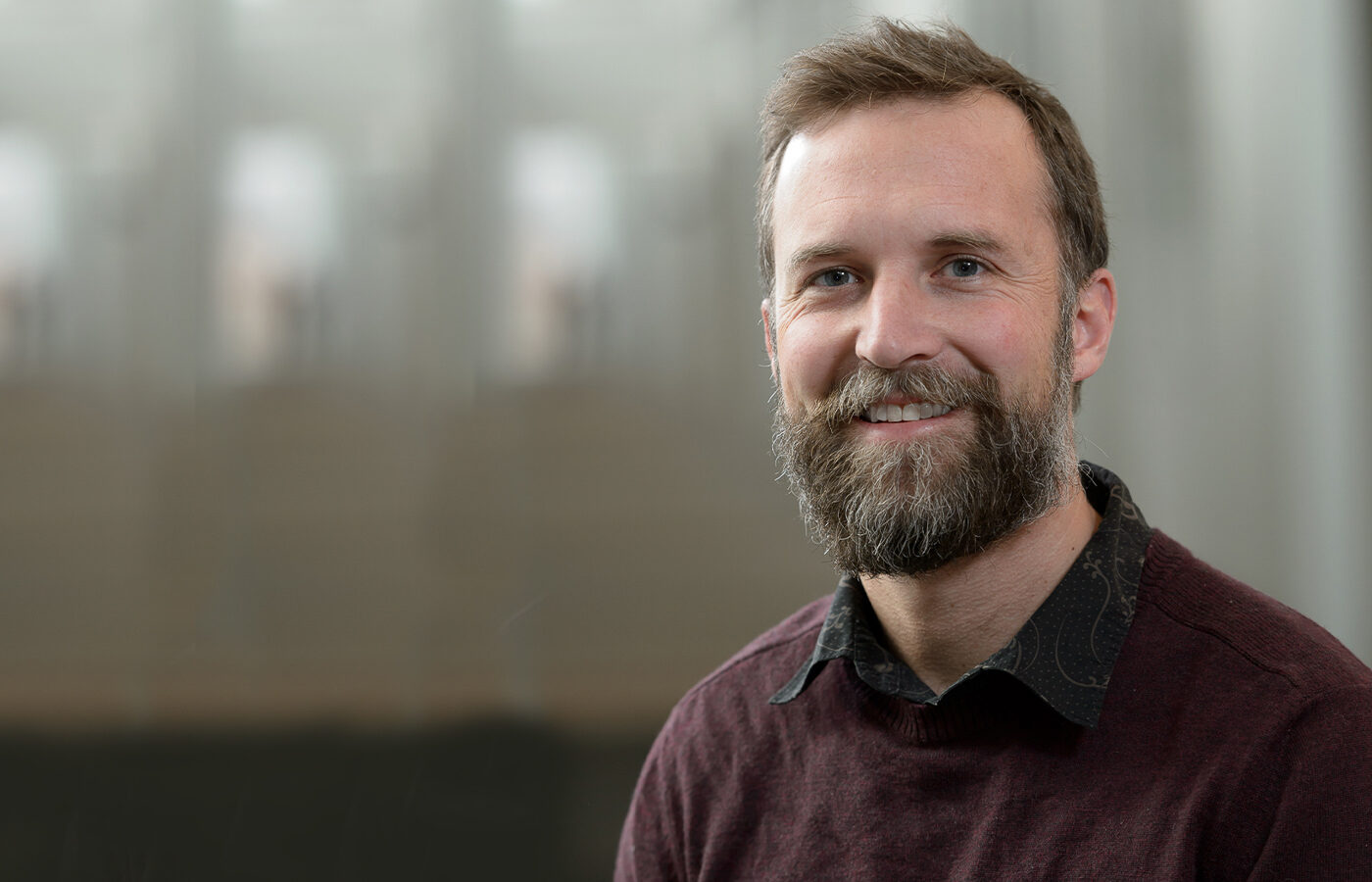 Person with light hair and a beard wearing a maroon sweater against a blurred gray background.
